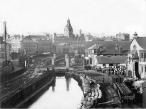 The Monmouthshire Canal at Mill Street 1914, in this view looking north from Mill Street, a barge navigates past the bascule bridge that provided access from the Mill Street railway yard to Cordes Dos Works on the left. It was around the time when regular canal traffic ceased to travel through to Moderator Wharf, Newport to discharge goods. Wagons to and from the works would have been shunted by the industrial locomotive seen in the distance between the arms of the bridge. Newport Reference Library